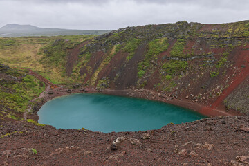 A rain-darkened rim above vivid blue water appears at Kerid Crater, Iceland. Diffuse afternoon light illuminates wet scoria and moss above the crater lake during rainy overcast conditions. © Artem