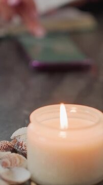 Close up of lit candle with seashells on dark surface soft warm light and shallow depth of field. Background includes blurred green book or tablet seashell