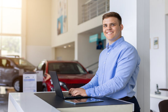 Smiling young car sales consultant working on laptop at dealership counter, representing professional automotive customer service, digital sales, and vehicle retail