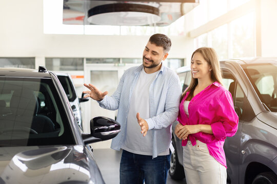 Young couple examining new vehicle at modern car dealership showroom while choosing automobile features before purchase decision