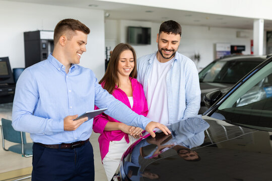 Salesman using digital tablet demonstrating car hood to young couple, discussing vehicle features and details in modern dealership showroom