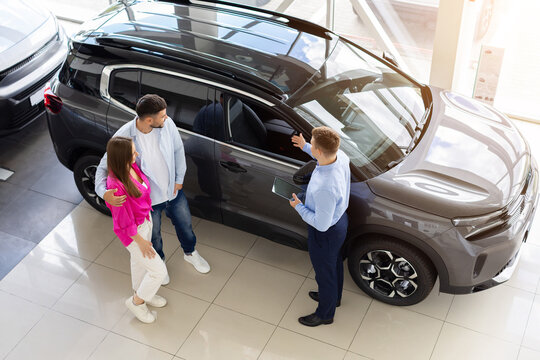 Young couple listening to car sales consultant beside SUV in modern dealership showroom while discussing vehicle features, specifications, and purchase options