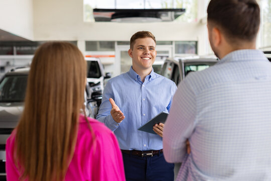 Car sales consultant speaking with young couple in bright dealership showroom, using a tablet while explaining vehicle details, pricing, financing, features, and purchase options