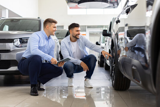 Salesman and customer crouching to inspect car tire and wheel closely together, two males comparing vehicles in dealership showroom