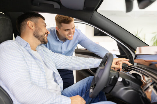 Car sales consultant demonstrating dashboard technology to young male buyer seated inside a showroom vehicle, explaining touchscreen controls, features, and driving functions