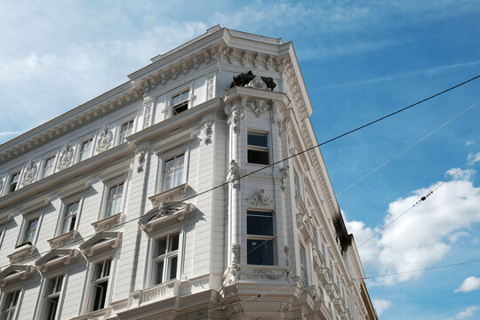 An elegant ornate corner facade of a historic white apartment building featuring classic decorative reliefs and bay window under a bright blue sky with clouds in Vienna, Austria.