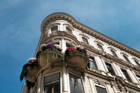 An elegant ornate corner facade of a historic apartment building featuring decorative balconies adorned with vibrant purple and white flowers under a clear blue sky in Vienna, Austria.