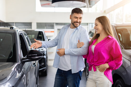 Young couple examining new car together in dealership showroom, smiling and discussing exterior design, vehicle features, quality, and options before making purchase decision