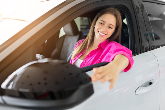 Smiling young woman examining side mirror while seated in showroom car, checking exterior design, visibility, comfort, and vehicle features before making a purchase decision