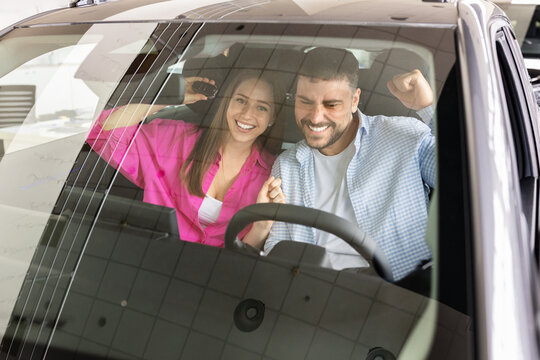 Excited young couple celebrating inside new car at dealership showroom, smiling through the windshield while proudly holding the vehicle key after successful purchase