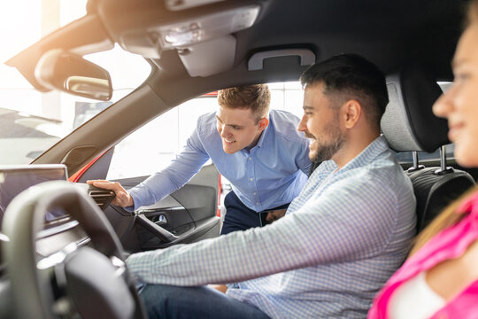 Car sales consultant explaining dashboard controls and interior features to young couple seated inside a showroom vehicle, helping them evaluate technology and purchase options