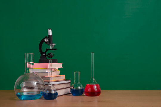 School background, chemistry class concept. Microscope, textbooks and chemistry glassware with colored solution in front of chalkboard. Empty space, copy space.