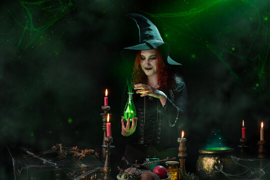 Witch conjuring a love potion, cooking up love, casts a spell, bottle with glowing poison in her hands. Young woman, a magician girl with red hair, dressed in Halloween attire, wearing conical hat.