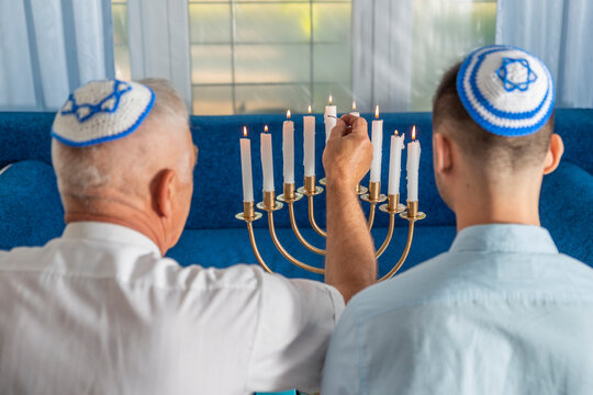 Hanukkah holiday. A senior man and young guy wear kippahs on heads in front of Jewish Hanukkah Menorah, lighting fire of candles. Yarmulke with Star of David. Traditional Hebrew Festival of Lights