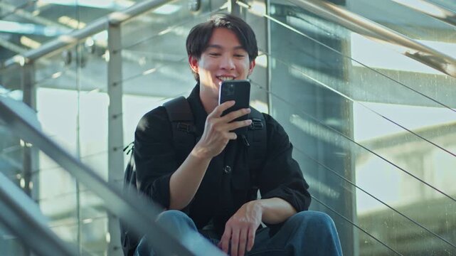 Happy young Asian man using smartphone while sitting on stairs in modern urban building, smiling male traveler with backpack checking mobile app, online communication, travel technology