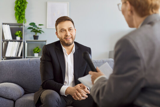 Businessman in interview speaks into microphone with journalist. He has focused conversation with interviewer in office. Woman interviewing guest. Concept media communication and corporate dialogue.