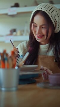 Happy young Asian woman painting pottery with brush in creative ceramic workshop, smiling female artist decorating handmade plate at craft studio, art hobby, creativity, handmade design and leisure ac