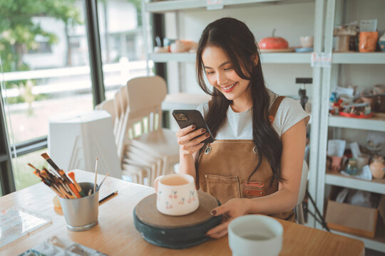 Smiling young woman using smartphone while working in pottery studio with handmade ceramic mug, creative female artisan enjoying art, craft, technology and modern digital lifestyle in bright workshop