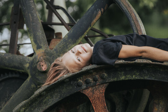 Young child with long hair lying on a large, rusty gear of an old machine surrounded by greenery in an outdoor setting