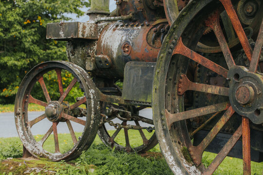 Vintage steam engine with rusted metal components and wooden wheels displayed on grassy area, surrounded by trees and a clear sky in the background