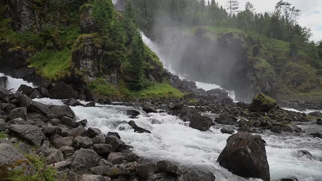 Misty waterfall cascades through mossy cliffs and a boulder-strewn riverbed in Norway during summer.