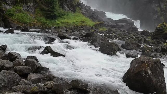 Turbulent whitewater cascades over dark boulders through a misty mountain landscape surrounded by lush green vegetation.
