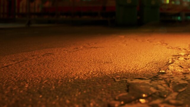 A view of water pooling on the ground at night. The location shows a parking area with lights in the background. The surface has a wet appearance reflecting nearby lights.