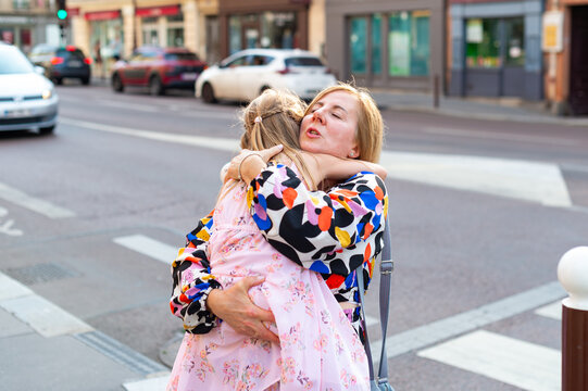 A mother holds and hugs her young daughter on a city sidewalk in Paris, France. Vehicles and storefronts are visible along the street in the background.