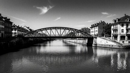 Fototapeta premium Black, white view of metal arch bridge over river in European city. Old buildings line canal banks near urban waterway. Calm water reflects bridge structure, architecture under cloudy sky. Scenic