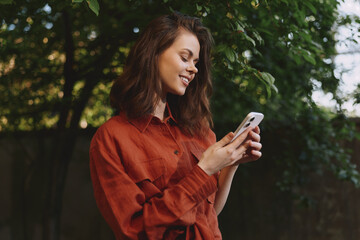 Naklejka premium Woman lifestyle shot outdoors wearing a red shirt, smiling while using a smartphone, natural light, green trees in the background, relaxed candid moment in a park or garden setting