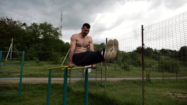 Strong man trains abdominal muscles raising legs on parallel bars at sports ground. Young shirtless sportsman exercising at nature. Sporty athlete working out at nature. Concept of sportive lifestyle