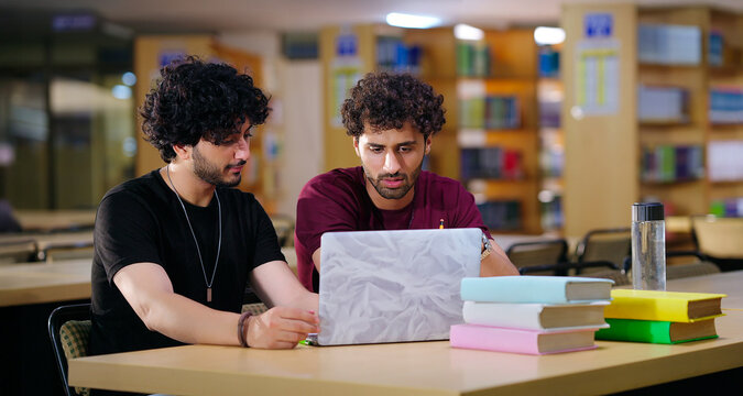 Indian young adult male student sitting on desk table use laptop look screen check new exam notes e work gen z boys man guy do study talk share task plan ideas learn course skill indoor library place