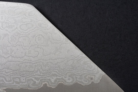 Extreme close up of a sharp textured Damascus steel knife blade tip showcasing organic wavy layered patterns placed over a black felt surface.