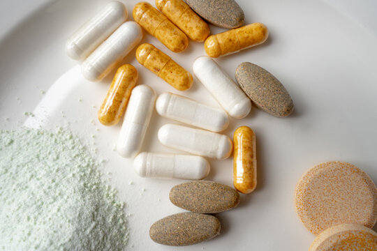Macro close-up of white and yellow gelatin supplement capsules, herbal tablets and green superfood powder on a white ceramic plate