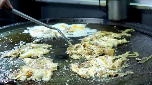 Thai street food vendor flips a crispy fried mussel pancake on a hot griddle at a night market, capturing authentic seafood preparation and local dining culture for food and travel content.