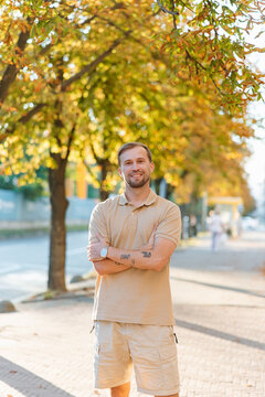 Handsome smiling young man standing outdoor on city street against autumn tree background and looking at camera