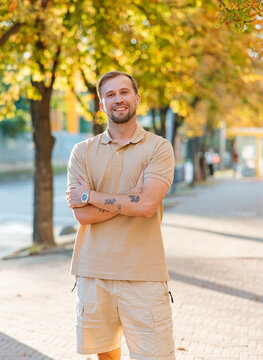 Handsome smiling young man standing outdoor on city street against autumn tree background and looking at camera