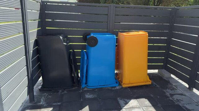Three colorful recycling bins in black, blue, and yellow in an outdoor waste enclosure with dark slatted fence, waste management, residential area