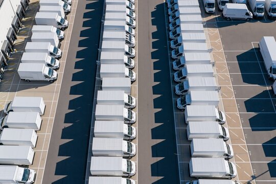 Aerial drone view above rows of identical white delivery vans parked in precise parallel lines within a large commercial vehicle depot, casting repeating dark shadows under bright morning sunlight