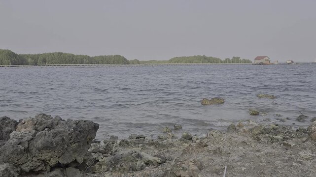 Rocky coastline with volcanic stone and calm ocean water against distant green shore and small house for travel and environmental design context
