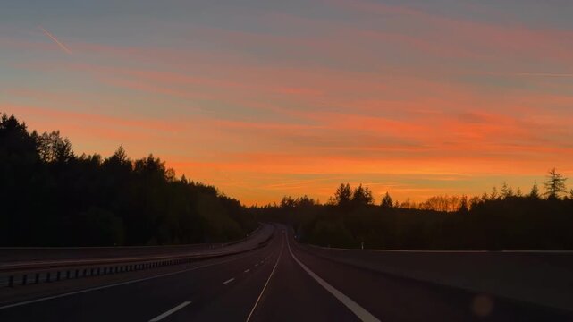 A smooth driving POV shot through a car windshield while traveling fast along an open highway autobahn toward a spectacular colorful sunset. Golden, orange, and purple sky reflections.