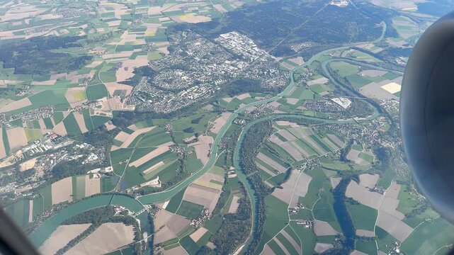 A scenic high-angle video captured through an airplane passenger window, overlooking the picturesque Austrian countryside. The aerial shot reveals neatly divided green agricultural fields, winding