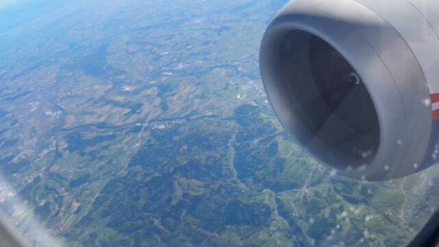 A scenic high-angle video captured through an airplane passenger window, overlooking the picturesque Austrian countryside. The aerial shot reveals neatly divided green agricultural fields, winding