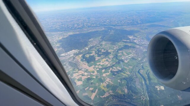 A scenic high-angle video captured through an airplane passenger window, overlooking the picturesque Austrian countryside. The aerial shot reveals neatly divided green agricultural fields, winding