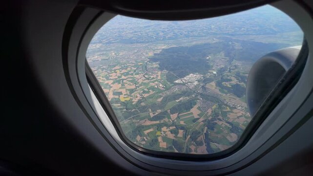 A scenic high-angle video captured through an airplane passenger window, overlooking the picturesque Austrian countryside. The aerial shot reveals neatly divided green agricultural fields, winding