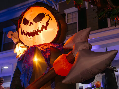 A giant inflatable pumpkin monster stands outside a decorated home on Halloween night. Bright internal lighting highlights the spooky face and oversized hands.