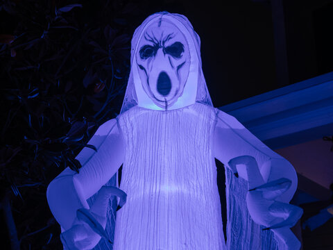 A glowing ghost inflatable with a haunting face stands outside during Halloween evening. Blue and purple lighting adds a dramatic spooky atmosphere.