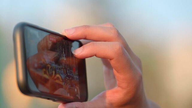 Hand holding smartphone in landscape orientation with fingertip touching glowing screen, showing blurred outdoor scene and playback interface under warm sunset light, shallow depth of field