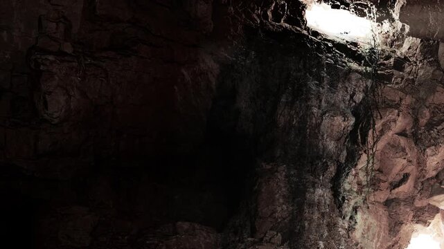 Inside a dark cave, a beam of light shines through a small opening above. The rocky walls show shadows and textures from the surrounding stone. It appears to be early morning.