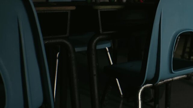 A classroom is empty, showing blue chairs and wooden desks. The light falls softly through the window, creating shadows. The scene is quiet and still, suggesting a break in activity.